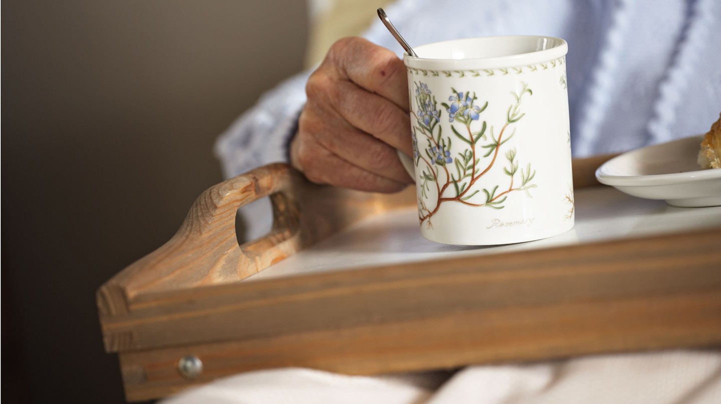 wdv-340961 Häusliche Pflege mit Heilpflanzen und Hausmitteln: Fotoausschnitt eines Pflegebedürftigen, der im Bett ein Tablett mit einer großen Tasse Kräutertee hält.